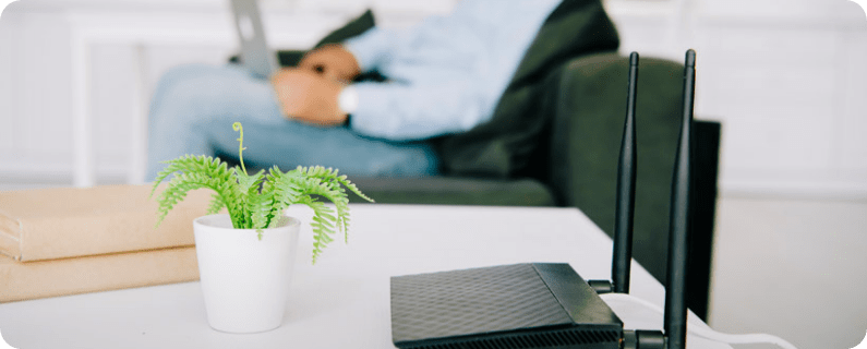 White coffee table with a potted fern, two books, and a Wi‑Fi router; blurred person using a laptop on a sofa in the background.